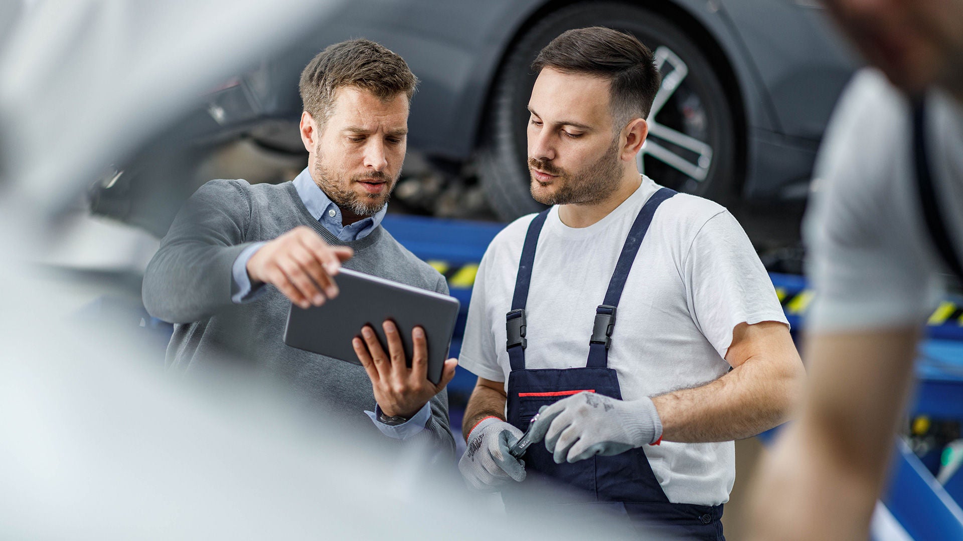 Mechanic in overalls discussing with a man holding a tablet inside an auto repair shop.