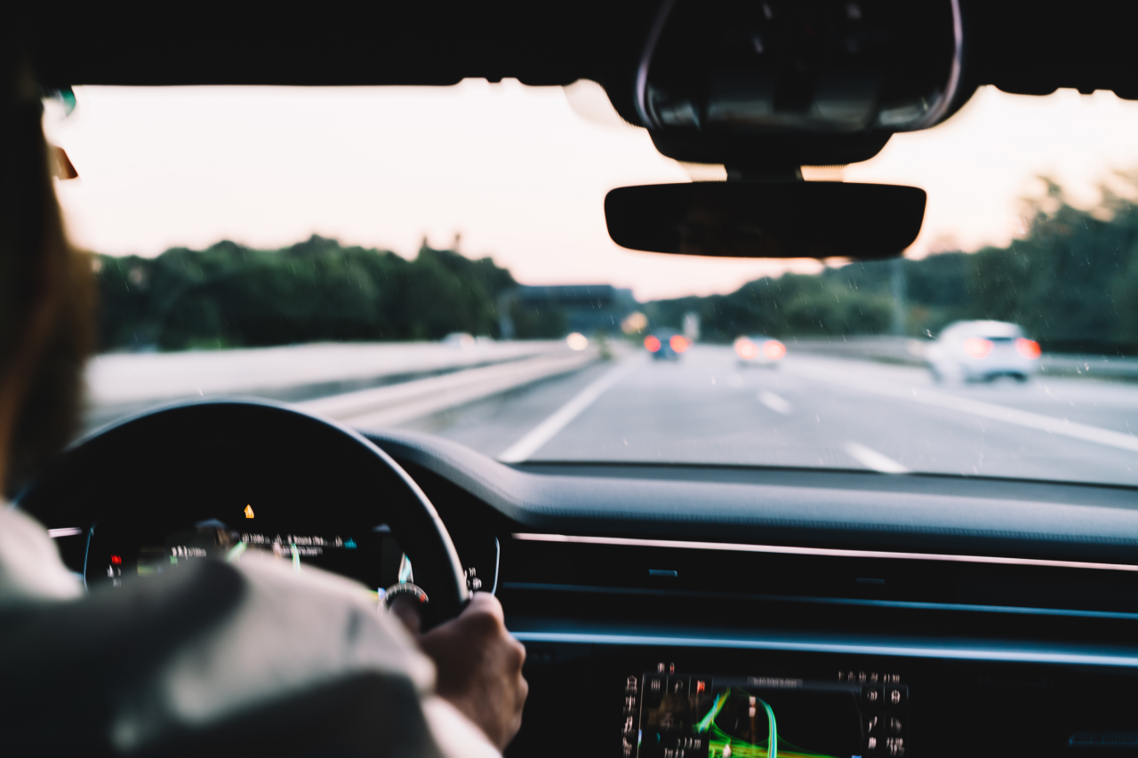 Blurred view from inside a car, showing a person driving on a highway with several cars visible ahead. The dashboard and steering wheel are in focus, while the road and surrounding greenery are out of focus in the background.